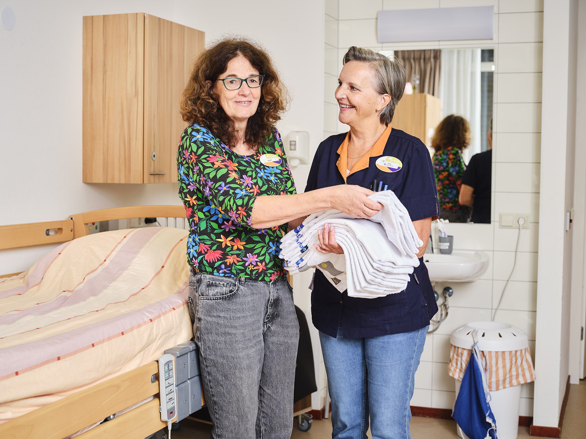 Twee medewerkers van Archipel houden schone handdoeken vast in de slaapkamer van een cliënt in een verzorgingshuis.