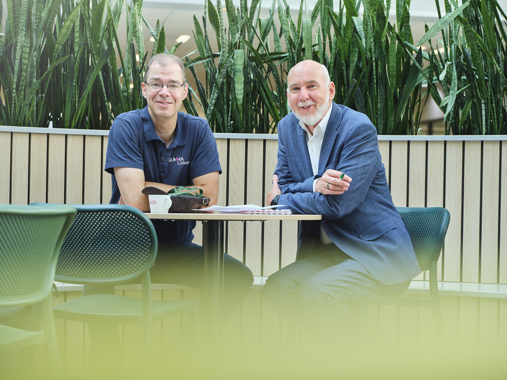 Een medewerker van Summa college zit samen met hoofd Facilitair Fred Wichard van Summa college aan tafel in een aula.