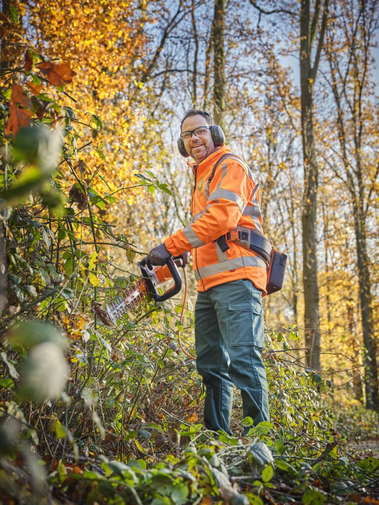 Een medewerker van Ergon Groen snoeit takken vol gele en oranje herfstbladeren met een heggenschaar.