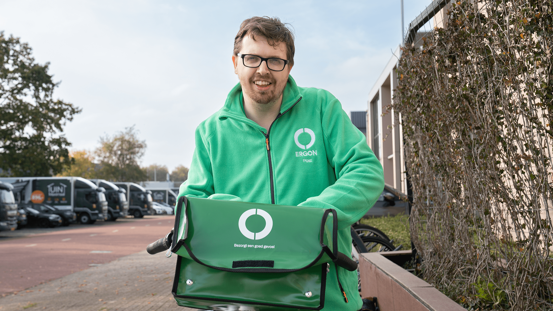 Een medewerker van Ergon Post staat op een parkeerplaats. Hij zit op zijn bezorgfiets en lacht.