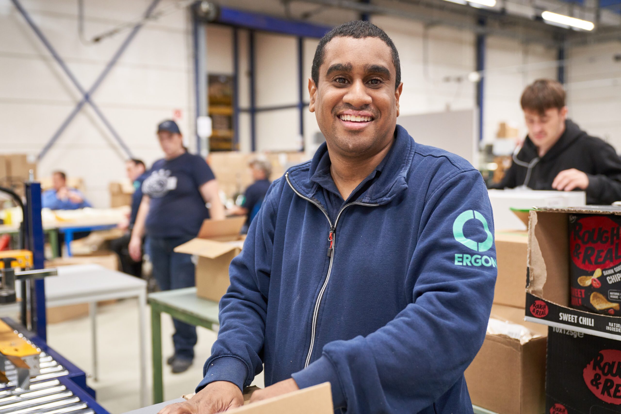 Een medewerker van Ergon Productie vouwt een doos. Op de achtergrond zijn zijn collega's ook in- om- en verpakwerkzaamheden aan het uitvoeren.