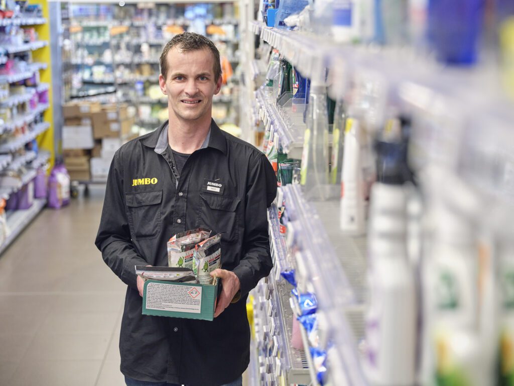 Een medewerker van Jumbo staat in een gangpad van de supermarkt met enkele dozen schoonmaakartikelen in zijn handen.