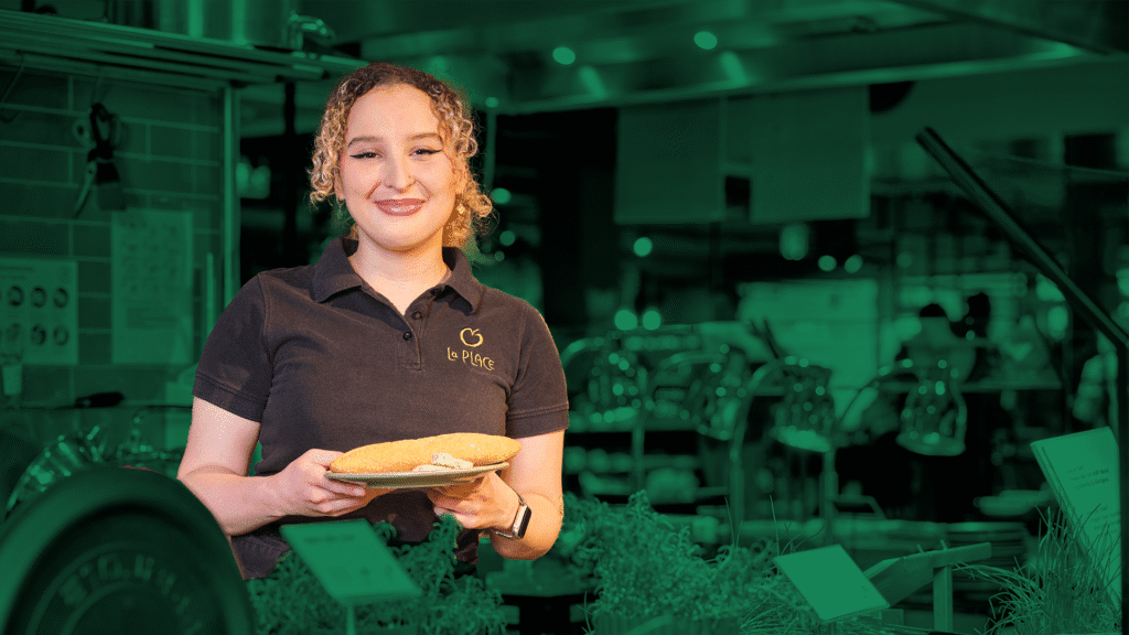 Een medewerker van La Place staat met een bordje waarop een broodje ligt trots achter een toonbank vol etenswaren.