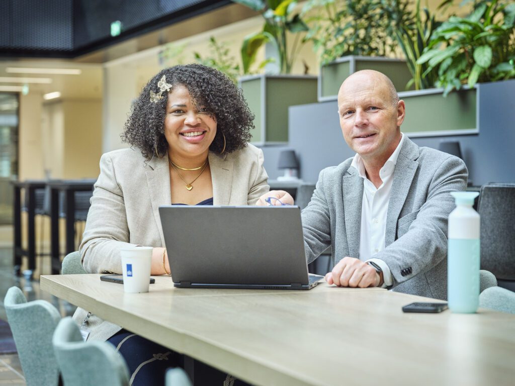 Een leidinggevende van de Belastingdienst zit met een gedetacheerde medewerker aan een bureau. Ze kijken allebei in de camera.